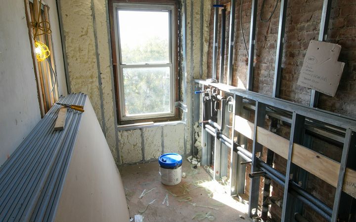 Older room stripped to studs and insulation, with a drywall sheet leaning by the window